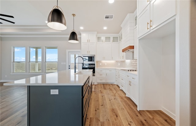 kitchen featuring stainless steel microwave, hanging light fixtures, a kitchen island with sink, light countertops, and white cabinetry