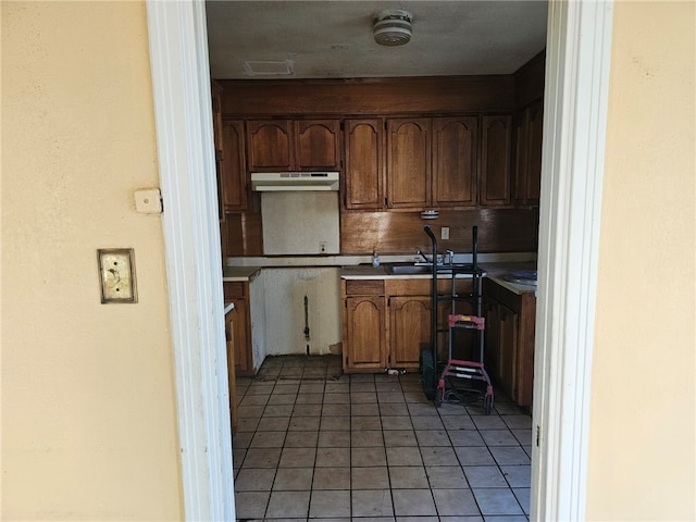 kitchen with light tile patterned flooring, a sink, light countertops, under cabinet range hood, and brown cabinets