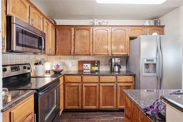 kitchen featuring stainless steel appliances, backsplash, dark stone counters, brown cabinetry, and dark wood finished floors