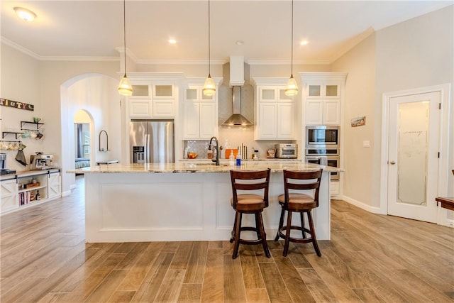 kitchen featuring light stone counters, a center island with sink, hanging light fixtures, stainless steel appliances, and wall chimney range hood