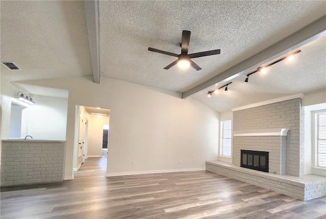 unfurnished living room featuring wood-type flooring, lofted ceiling with beams, ceiling fan, a brick fireplace, and a textured ceiling