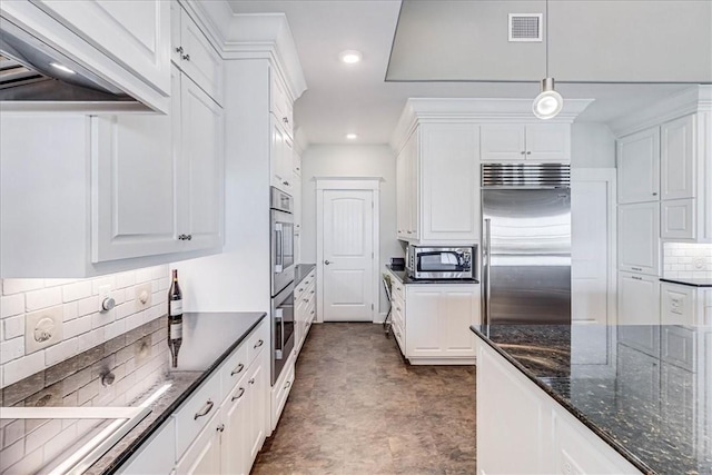 kitchen featuring stainless steel appliances, dark stone countertops, white cabinetry, hanging light fixtures, and range hood