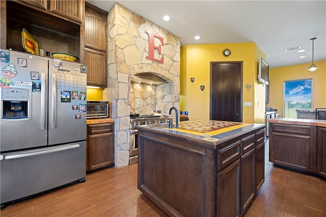 kitchen featuring stainless steel appliances, dark brown cabinets, dark hardwood / wood-style floors, hanging light fixtures, and an island with sink