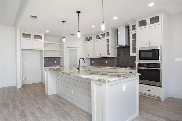 kitchen featuring pendant lighting, white cabinets, wall chimney range hood, an island with sink, and stainless steel appliances