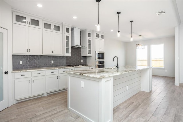 kitchen with stainless steel appliances, a kitchen island with sink, wall chimney range hood, white cabinets, and hanging light fixtures