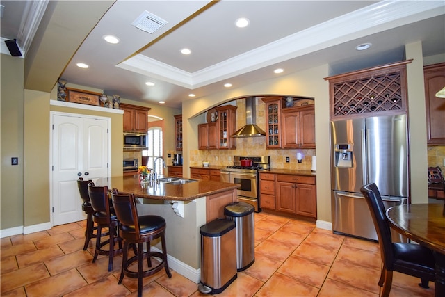 kitchen featuring sink, stainless steel appliances, wall chimney range hood, backsplash, and a center island with sink