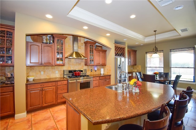 kitchen with wall chimney range hood, a raised ceiling, decorative light fixtures, a kitchen island with sink, and appliances with stainless steel finishes