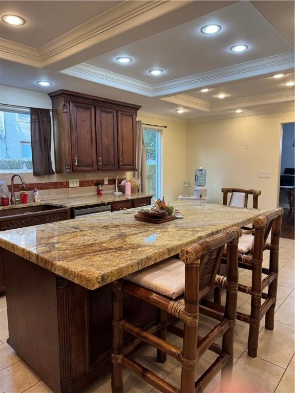 kitchen featuring a sink, a tray ceiling, a breakfast bar, light stone counters, and ornamental molding
