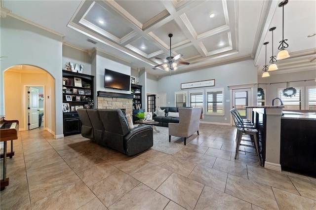 living room featuring a high ceiling, coffered ceiling, a stone fireplace, ceiling fan, and ornamental molding