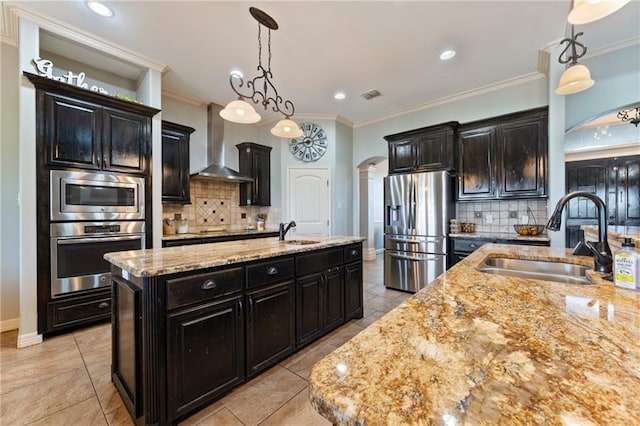 kitchen featuring sink, stainless steel appliances, wall chimney range hood, decorative light fixtures, and a center island with sink