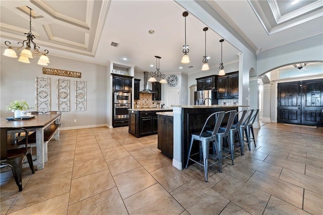 kitchen with appliances with stainless steel finishes, tasteful backsplash, wall chimney range hood, pendant lighting, and a center island with sink