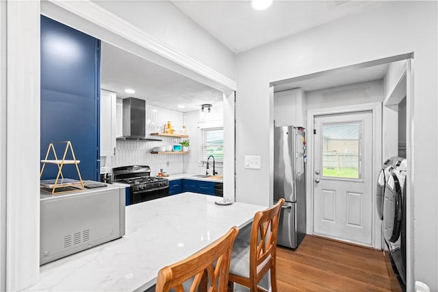 kitchen with gas range, freestanding refrigerator, wall chimney range hood, washing machine and dryer, and blue cabinetry