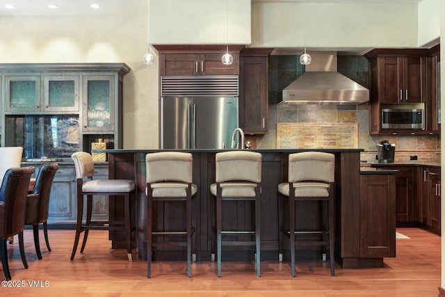 153 Village Walk, Beaver Creek, CO 81620 - kitchen with wall chimney range hood, appliances with stainless steel finishes, dark countertops, light wood-style floors, and decorative backsplash