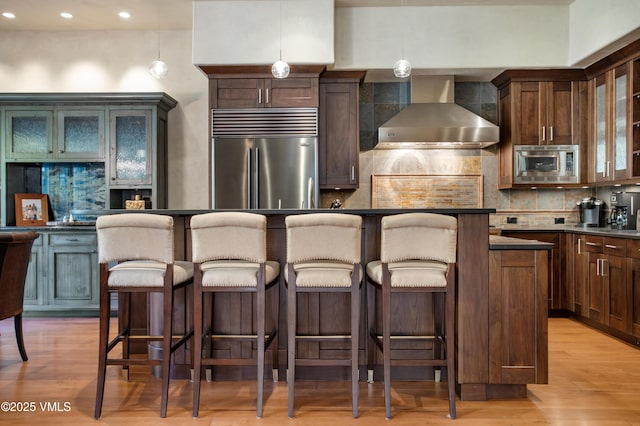 kitchen with a kitchen island, a breakfast bar, hanging light fixtures, built in appliances, and wall chimney range hood