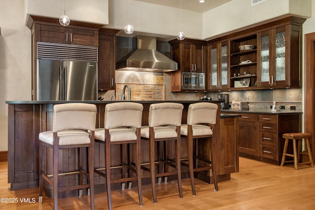 bar with tasteful backsplash, built in appliances, hanging light fixtures, light hardwood / wood-style flooring, and wall chimney range hood