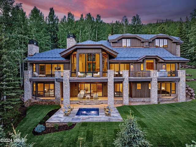 rear view of property featuring a lawn, a patio, a balcony, stone siding, and a chimney