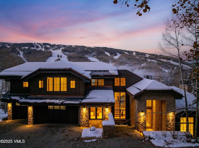 snow covered property featuring stone siding, decorative driveway, an attached garage, and a mountain view