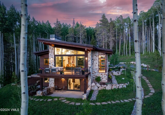 back of house at dusk with stone siding, outdoor lounge area, a patio, and a lawn