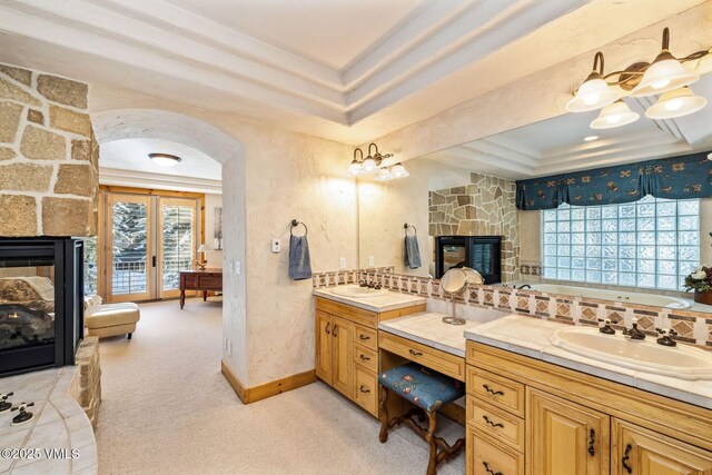 bathroom with a tray ceiling, vanity, a stone fireplace, and a tub