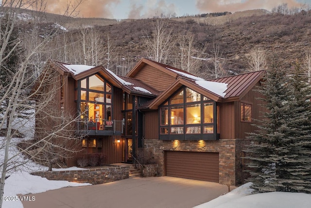 rustic home featuring a standing seam roof, metal roof, a garage, stone siding, and driveway