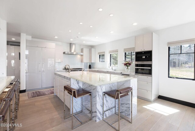 kitchen with wall chimney exhaust hood, a kitchen bar, sink, a kitchen island with sink, and white cabinets