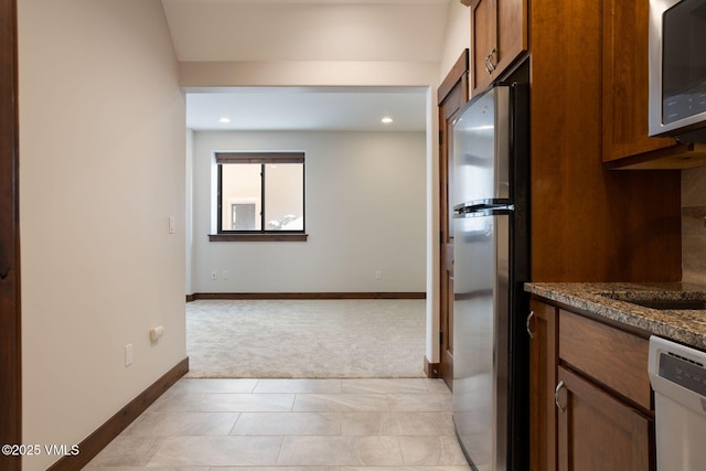 kitchen featuring light carpet, baseboards, brown cabinetry, dark stone countertops, and stainless steel appliances