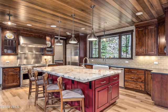 kitchen featuring decorative light fixtures, a center island with sink, dark stone countertops, premium appliances, and wall chimney range hood