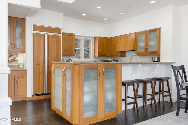 kitchen with a breakfast bar area, brown cabinetry, recessed lighting, dark wood-type flooring, and glass insert cabinets