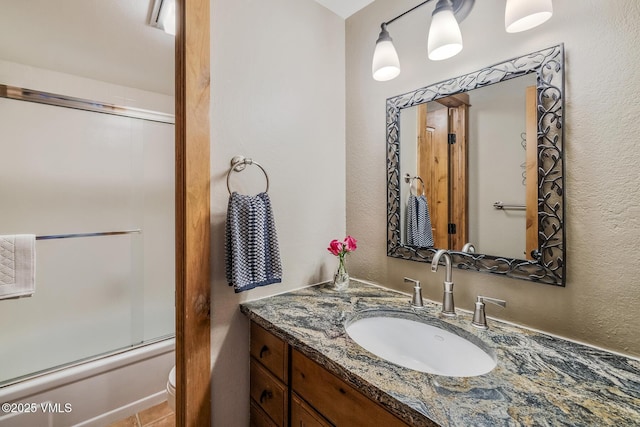 bathroom with combined bath / shower with glass door, toilet, vanity, and a textured wall