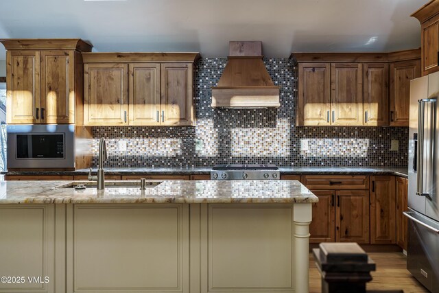 kitchen with high end fridge, sink, dark stone countertops, a kitchen island with sink, and backsplash