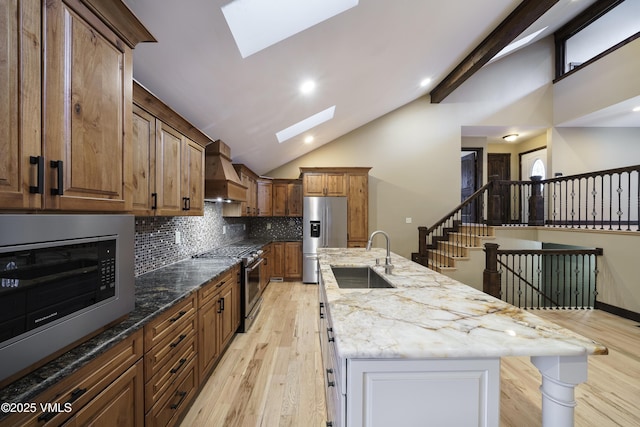 kitchen with sink, appliances with stainless steel finishes, a skylight, custom exhaust hood, and a large island with sink