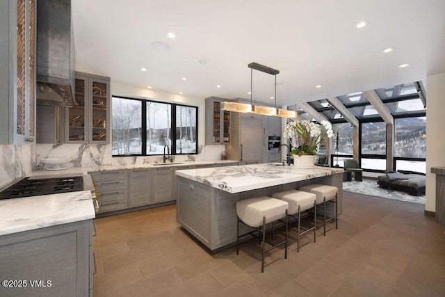 kitchen with gray cabinetry, a sink, exhaust hood, decorative backsplash, and a kitchen bar