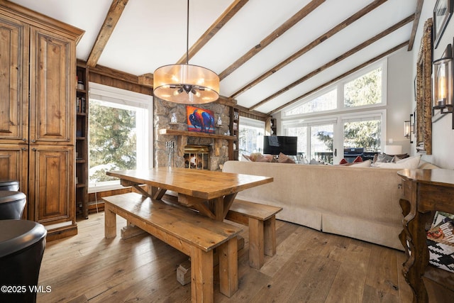 dining space featuring beam ceiling, hardwood / wood-style floors, a stone fireplace, and a chandelier