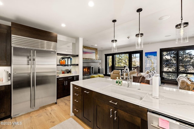 kitchen with a warm lit fireplace, dark brown cabinetry, dishwasher, open floor plan, and stainless steel built in refrigerator