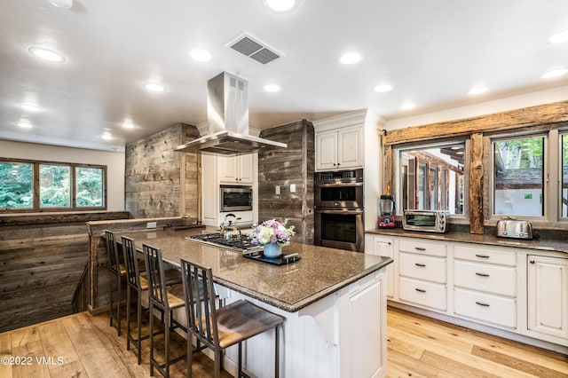 kitchen with appliances with stainless steel finishes, a breakfast bar area, white cabinets, dark stone counters, and island exhaust hood