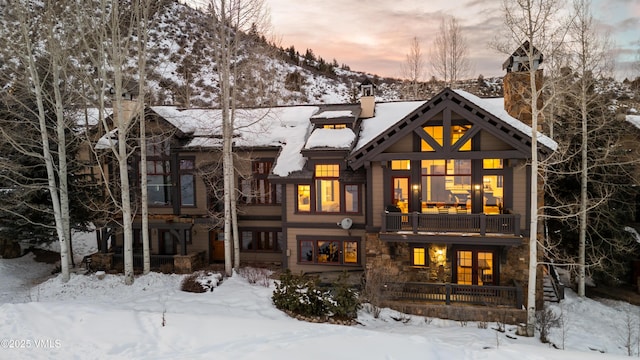 snow covered house featuring a balcony, stone siding, a chimney, stairway, and a mountain view