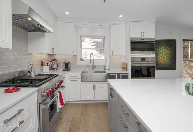 kitchen with white cabinetry, sink, stainless steel appliances, wall chimney range hood, and light hardwood / wood-style flooring