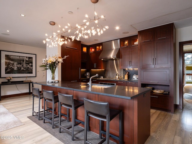 kitchen with glass insert cabinets, light wood-type flooring, a kitchen breakfast bar, wall chimney exhaust hood, and a sink