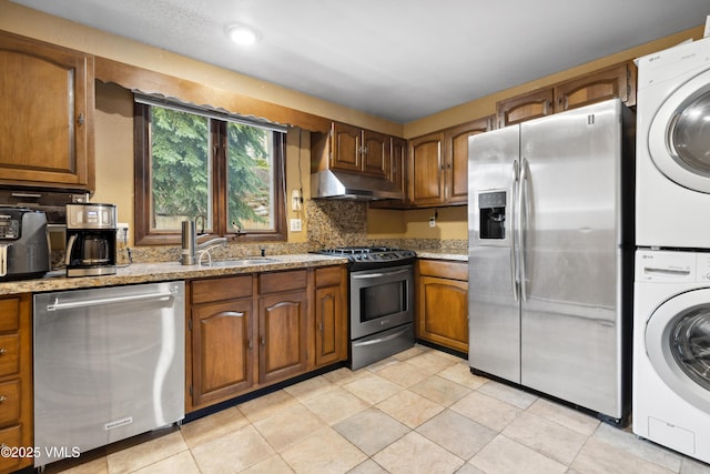 kitchen with brown cabinetry, stacked washer / dryer, under cabinet range hood, a sink, and appliances with stainless steel finishes