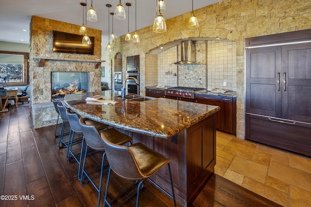 kitchen with appliances with stainless steel finishes, decorative light fixtures, sink, a large island with sink, and wall chimney range hood