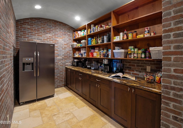 bar featuring dark brown cabinets, stainless steel refrigerator with ice dispenser, brick wall, vaulted ceiling, and dark stone counters