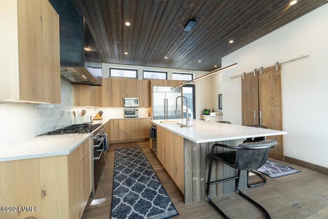 kitchen featuring high end appliances, tasteful backsplash, a large island with sink, a barn door, and wall chimney range hood