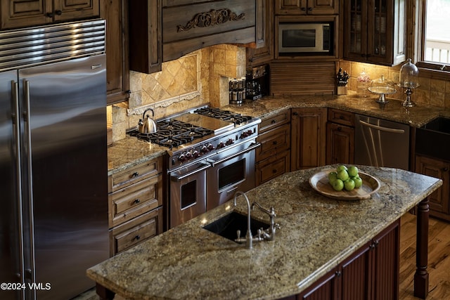 kitchen featuring high end appliances, an island with sink, a sink, stone counters, and backsplash