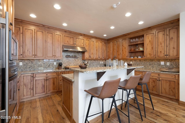 kitchen with an island with sink, light stone counters, a breakfast bar, under cabinet range hood, and open shelves