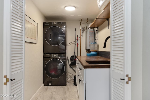 clothes washing area with stacked washing machine and dryer, light marble finish flooring, cabinet space, water heater, and baseboards