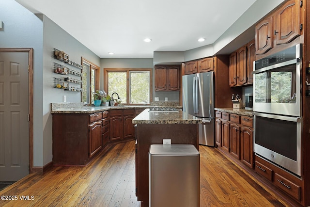 kitchen with stainless steel appliances, hardwood / wood-style flooring, a kitchen island, a sink, and light stone counters