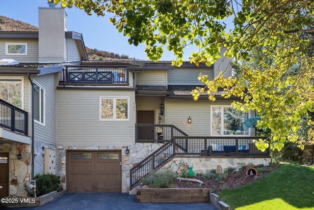 view of front of home with a balcony, an attached garage, a chimney, stone siding, and asphalt driveway