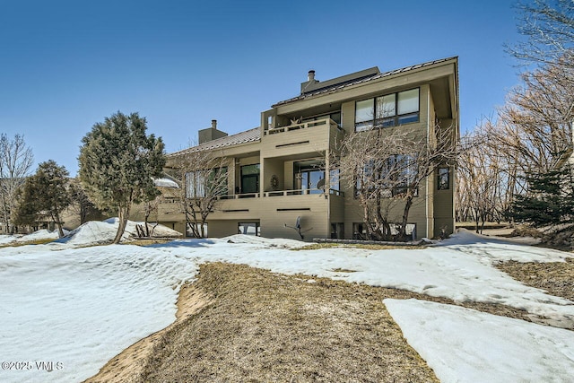 snow covered back of property featuring a standing seam roof, a balcony, metal roof, and a chimney