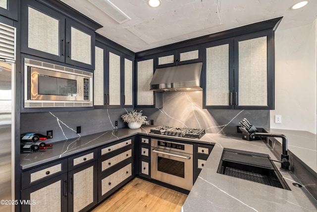 kitchen featuring light wood-style flooring, a sink, decorative backsplash, under cabinet range hood, and appliances with stainless steel finishes