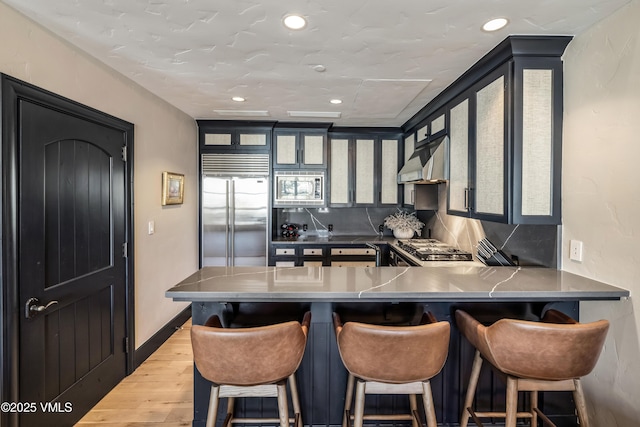 kitchen with under cabinet range hood, wood finished floors, a peninsula, glass insert cabinets, and built in appliances